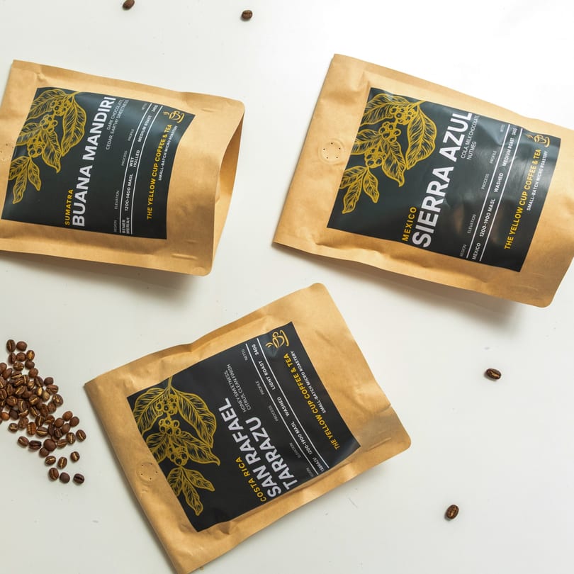 Three bags of "The Yellow Cup Coffee & Tea" coffee beans with scattered coffee beans.