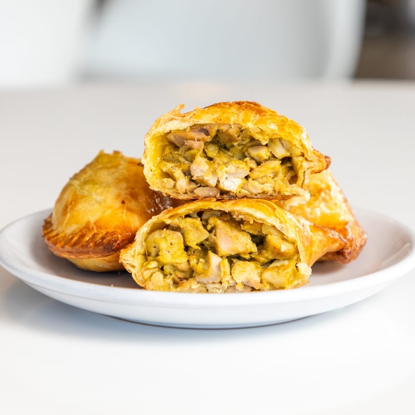 Golden-brown empanadas, one cut open to reveal a savory chicken and vegetable filling, on a white plate.
