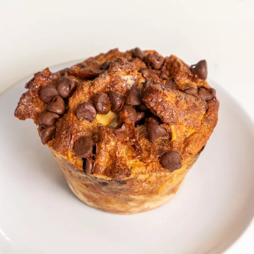 A golden-brown muffin, generously topped with dark chocolate chips, sits on a white plate.