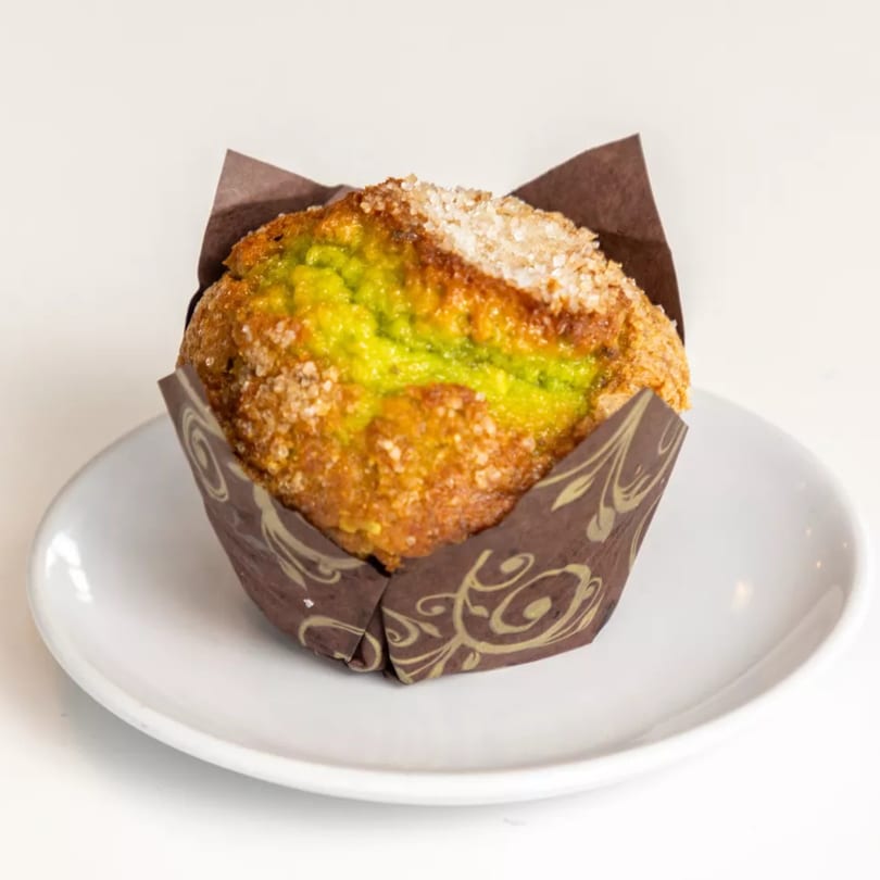 A pistachio muffin with a sugar topping in a brown wrapper on a white plate.